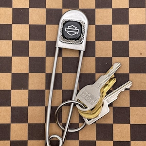 Pode incluir: Um grande alfinete de segurança prateado com um logotipo Harley-Davidson preto. O alfinete contém um porta-chaves com várias chaves. O fundo é um padrão xadrez castanho e preto.