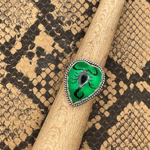May include: A heart-shaped ring with a green center featuring a black scorpion design. The ring has a silver-colored, twisted rope-like border. The ring is displayed on a wooden dowel against a snakeskin pattern background.