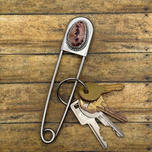 Pode incluir: Um porta-chaves de alfinete de ama grande em metal prateado com uma pedra decorativa oval de padrões marmoreados castanhos e brancos, engastada numa borda de metal torcido. Um anel de metal está preso ao alfinete e segura três chaves.