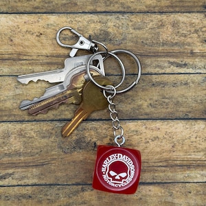 Pode incluir: Um porta-chaves de metal com várias chaves de prata e latão, apresentando um pendente vermelho em forma de cubo com o logótipo branco Harley-Davidson Motorcycles e um gráfico de caveira impresso na frente, preso por uma corrente curta.