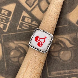 May include: A square ring with a white center featuring a red design. The ring has a silver-colored, twisted rope-like border. The ring is displayed on a wooden dowel against a patterned background.