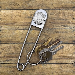 Pode incluir: Um alfinete de segurança grande em metal prateado com uma moeda americana de cinco centavos com a imagem de um bisão incrustada no topo. Um porta-chaves circular de metal está preso à base, segurando um conjunto de quatro chaves de metal.