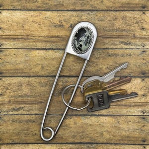Pode incluir: Um alfinete de segurança grande em metal prateado com um cabochão oval de pedra com padrão manchado em preto e verde. Um anel metálico circular está preso ao alfinete, segurando três chaves de metal variadas em tons de latão e prata.