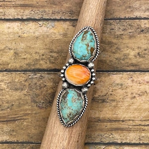May include: A silver ring featuring three stones: two turquoise teardrop-shaped stones and one orange oval stone. The stones are set in a silver setting with a twisted rope design around the edges. The ring is displayed on a wooden dowel.