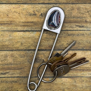 Pode incluir: Um porta-chaves grande de metal prateado em forma de alfinete de ama, com um cabochão em forma de lágrima com riscas multicoloridas em camadas de vermelho, azul, branco e preto. A pedra está engastada numa borda de metal torcido.
