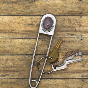Pode incluir: Um alfinete de segurança grande em metal prateado como porta-chaves, com uma pedra oval lisa de cor malva escuro engastada numa moldura decorativa com textura de corda. Um anel metálico prende um conjunto de três chaves de metal variadas.