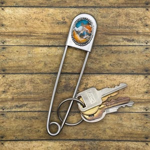 Pode incluir: Um porta-chaves grande de metal prateado em forma de alfinete de ama, com uma incrustação oval de pedra marmoreada em tons de turquesa, laranja e branco, rodeada por um rebordo entrançado. Três chaves metálicas estão presas por um anel.