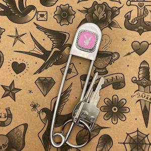 Könnte beinhalten: Eine silberne Sicherheitsnadel mit einem rosa und weißen Playboy-Hasen-Logo, die zwei Schlüssel an einem Ring hält. Die Nadel befindet sich vor einem braunen Hintergrund mit verschiedenen Tattoo-Illustrationen, darunter Vögel, Herzen und Blumen.