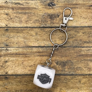 Pode incluir: Chaveiro de dados branco com o logotipo Harley-Davidson Cycles. O dado tem pontos pretos e é preso a uma corrente, argola e fecho de metal prateado. O fundo é uma superfície de madeira.