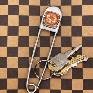 Pode incluir: Um porta-chaves de alfinete de segurança prateado grande com um logotipo Harley-Davidson com uma caveira em um fundo laranja. O porta-chaves contém várias chaves em um anel prateado. O fundo é um padrão xadrez marrom e bege.