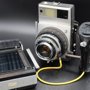 May include: A vintage silver and black large format camera with a lens, bellows, and a black grip. A film holder is to the left, and a yellow cable connects the camera to the grip. The camera is on a black surface.