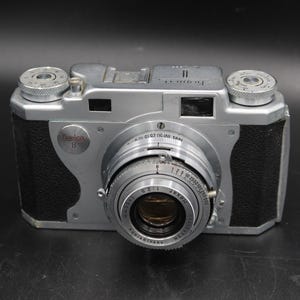 May include: A vintage silver and black camera with a prominent lens and dials. The camera has a classic design with black leather-like sides and a silver body. The lens has measurement markings. The camera is a Konica B model.