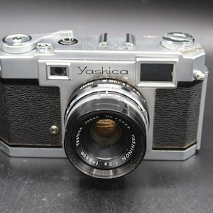 May include: A silver Yashica camera with a black lens and black textured grips. The lens is labeled "Yashinon 1:2.8 f=45mm". The camera has a retro design, with dials and buttons on top.