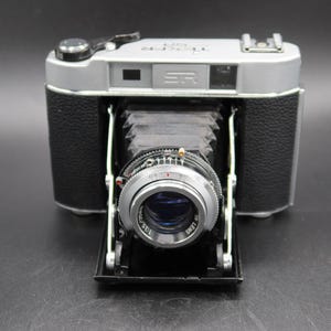 Op de afbeelding: Een vintage zilveren en zwarte SR-camera met een opvouwbare balg lens. De camera heeft een zilveren bovenkant en een zwarte kunstleren behuizing. De lens is zilverkleurig met een blauw interieur. De camera staat op een zwart oppervlak.