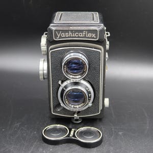 Könnte beinhalten: Schwarze Yashicaflex Zwei-Objektiv-Reflexkamera mit Markennamen oben. Die Kamera hat zwei Objektive, einen Fokusring und eine Objektivkappe. Die Kamera steht auf einer schwarzen Oberfläche.