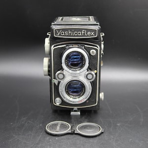May include: A black YashicaFlex twin-lens reflex camera with a silver Copal-MXV shutter assembly and two circular glass lenses. The camera body features textured black leatherette covering and includes a detached dual-lens cap at the base.