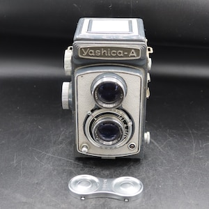May include: A vintage Yashica-A twin-lens reflex camera with a gray and silver finish. The camera has two lenses, a viewfinder, and a silver lens cap. The Yashica-A is a classic film camera.