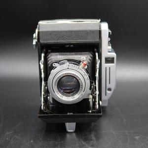 Könnte beinhalten: Eine Vintage-Faltkamera mit schwarzem Gehäuse und silbernen Metallakzenten. Die Objektiveinheit verfügt über einen silbernen Ring mit der Aufschrift 'Konishiroku Hexar 1:3.5 f=75mm' und ein Einstellrad für die Verschlusszeit. Mit Balgenmechanik.