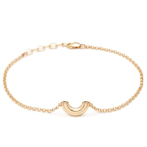 May include: A gold-colored bracelet featuring a curved, hollow design at the center. The bracelet has a delicate chain with an adjustable clasp. The central design has a rounded shape with two parallel lines.