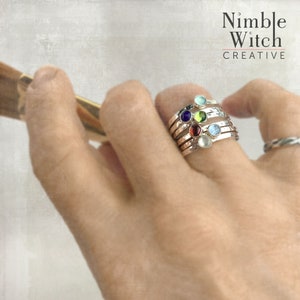 May include: Stackable silver rings with colorful gemstones, including blue, green, red, and purple. The rings are worn on a finger, showcasing the different colored stones. The rings are from Nimble Witch Creative.