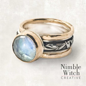 A blue flash moonstone in a round, facet cut  of 10mm sits in a gold filled setting on a sterling silver ring. This is the centerpiece of a 3-ring set with an antiqued, vintage style Florentine pattern. Ring is flanked by 2 sturdy gold filled rings.