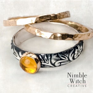 May include: Three rings: two gold-toned hammered bands and a silver band with a floral design and a yellow gemstone. The rings are stacked, with the silver ring at the bottom. The text "Nimble Witch Creative" is at the bottom.