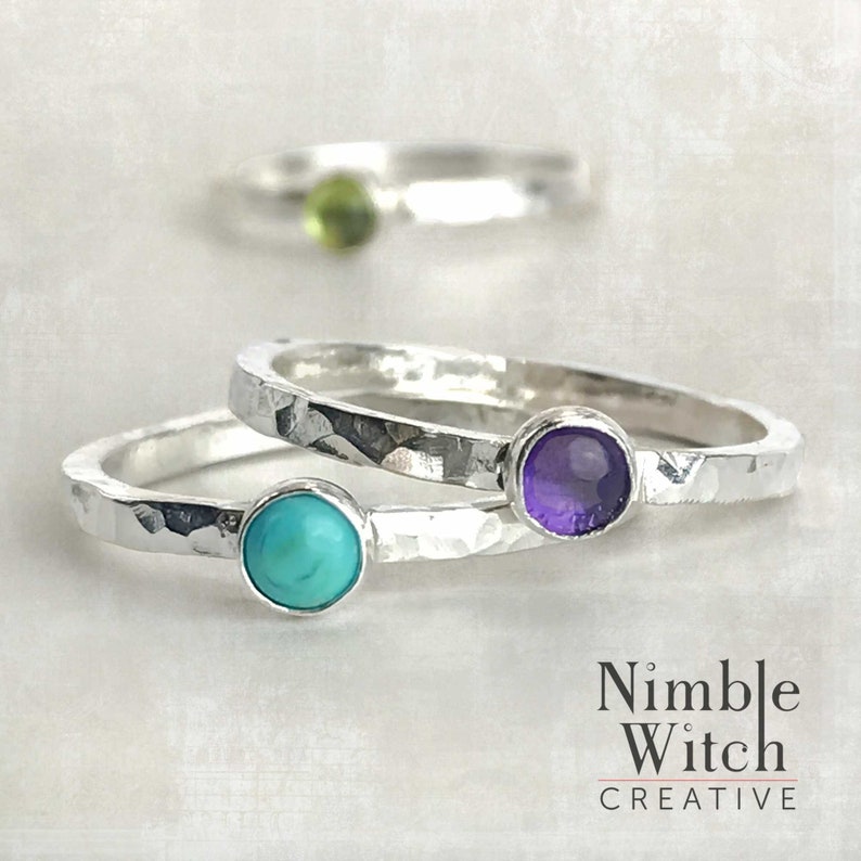 Three stacking natural birthstone rings. Each ring is set with a single stone in fine silver. Birthstones here are a Turquoise, Amethyst, and a Peridot which is shown in the background.