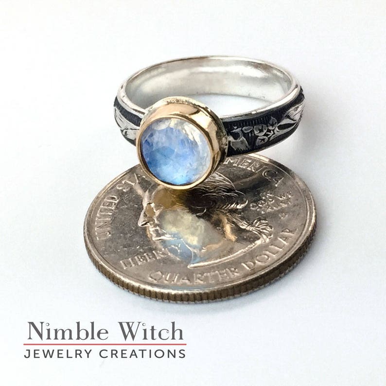 A blue flash moonstone in a round, facet cut  of 10mm sits in a gold filled setting on a sterling silver ring with an antiqued, vintage style Florentine pattern.