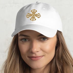 May include: White baseball cap with a gold-colored embroidered design on the front. The cap has a curved brim and a classic six-panel construction. The design appears to be a stylized emblem or symbol.