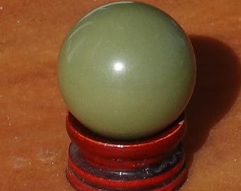 May include: A smooth, matte, olive green sphere resting securely on a small, circular, dark reddish-brown wooden stand. The sphere has a uniform, solid color and a polished surface, while the base features a tiered, carved design.