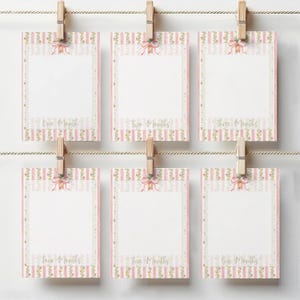 May include: Six baby milestone cards in pink and white, each clipped to a string with a wooden peg. The cards feature a pink striped border, floral details, and a bow. Each card is labelled with a month.