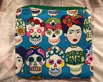 May include: A blue zippered pouch with a colorful pattern of sugar skulls and a Frida Kahlo-inspired face. The skulls are decorated with flowers, hearts, and other details.