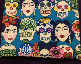 May include: A blue fabric pouch with a zipper closure. The pouch features a colorful pattern of sugar skulls and Frida Kahlo-inspired faces. The pattern includes flowers, hearts, and geometric designs.