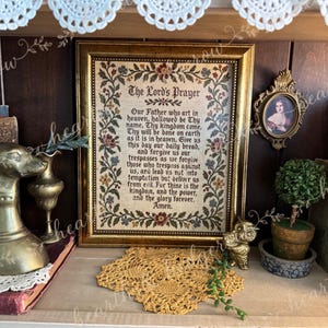 May include: A framed cross-stitch sampler featuring The Lord's Prayer in black text, surrounded by a floral border of red, blue, and green threads on a beige fabric background, enclosed in a rectangular gold-toned frame with a textured finish.