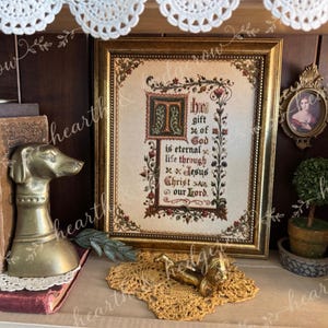 May include: A framed cross-stitch sampler featuring the text "The gift of God is eternal life through Jesus Christ our Lord" in a decorative border, displayed with a brass dog figurine, old books, a small portrait, and a topiary.