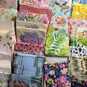 May include: A collection of square paper napkins featuring diverse printed patterns including floral arrangements, red gingham, strawberries, citrus slices, green tropical leaves, and a red wagon with American flags, arranged in a dense, overlapping display.