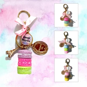 Könnte beinhalten: Ein rosa-goldener Schlüsselanhänger mit einem Anhänger, auf dem "Enni Norin" steht, und einem Stapel bunter Macarons. Der Schlüsselanhänger enthält außerdem einen goldenen Eiffelturm-Anhänger und einen goldenen Kreisanhänger mit einem Design.