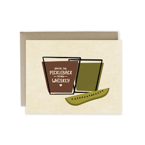 May include: A greeting card with a brown background and a green and brown illustration of two glasses. The text on the card reads "You're the pickleback to my whiskey".