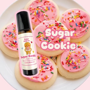 May include: A roll-on perfume bottle with a black cap and a label featuring a gingerbread man holding a cookie, placed in front of frosted sugar cookies with sprinkles. The text "Sugar Cookie" is also visible.