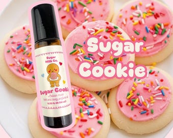 May include: A roll-on perfume bottle with a black cap and a label featuring a gingerbread man holding a cookie, placed in front of frosted sugar cookies with sprinkles. The text "Sugar Cookie" is also visible.
