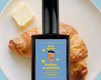 Könnte beinhalten: Eine Parfümflasche mit schwarzem Verschluss und einem rechteckigen Etikett mit der Aufschrift "Butter Croissant" von Sugar Milk Co. Das Etikett zeigt einen Cartoon-Bären und ist vor blauem Hintergrund platziert. Die Flasche steht auf einem weißen Teller mit einem Croissant und Butter.