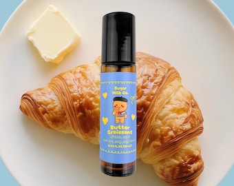 May include: A roll-on perfume bottle with a black cap and a blue label featuring a cartoon character and the text "Butter Croissant." The bottle sits on a white plate with a croissant and a pat of butter.