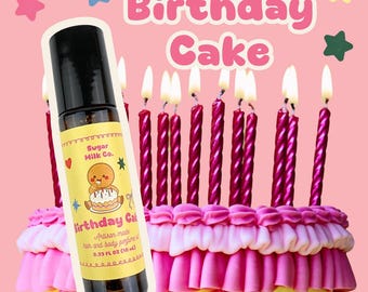 Op de afbeelding: Een roll-on flesje van het ambachtelijke parfum "Birthday Cake" wordt getoond voor een versierde taart met brandende kaarsen. De fles heeft een geel etiket met een gingerbread man afbeelding. De taart is geel met roze glazuur.