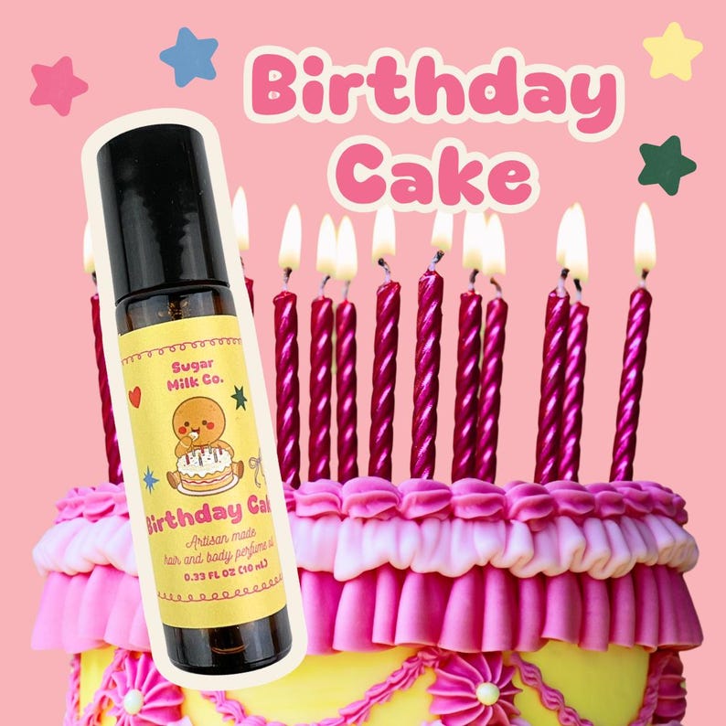 May include: A roll-on bottle of "Birthday Cake" artisan perfume is displayed in front of a decorated cake with lit candles. The bottle has a yellow label with a gingerbread man graphic. The cake is yellow with pink frosting and decorative elements.