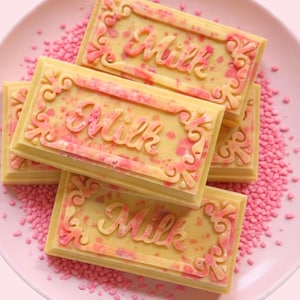 May include: A close-up of several rectangular milk chocolate bars with pink accents and the word "Milk" embossed on each. The bars are arranged on a pink plate with pink sprinkles.