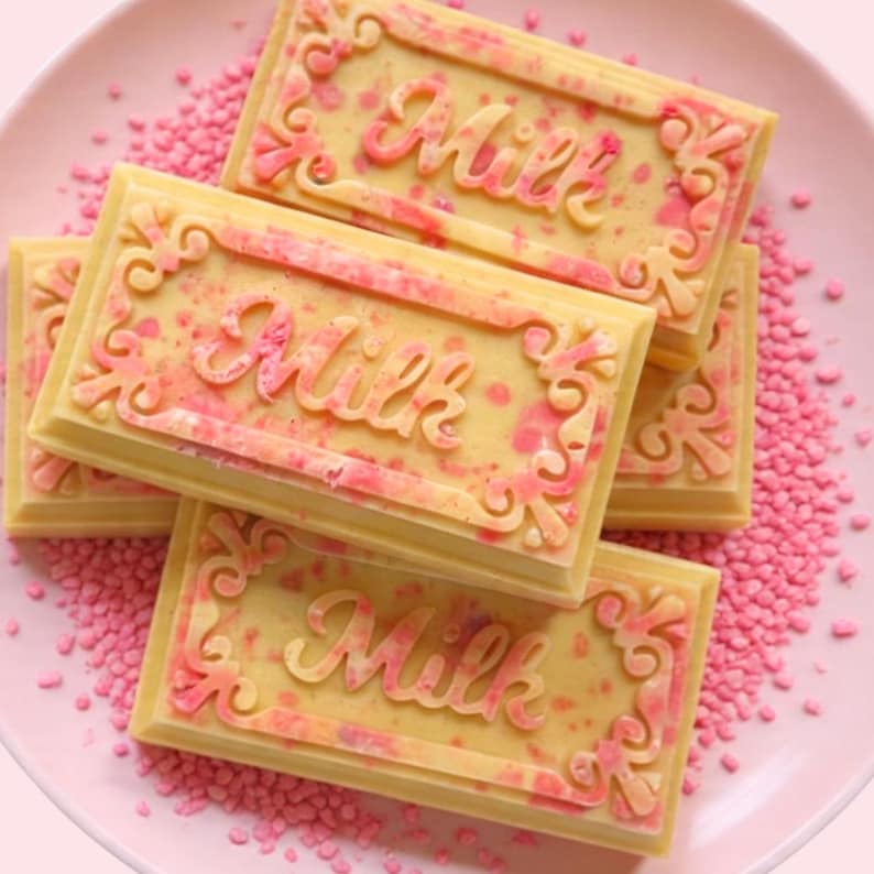 May include: A close-up of several rectangular milk chocolate bars with pink accents and the word "Milk" embossed on each. The bars are arranged on a pink plate with pink sprinkles.