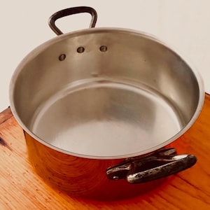 May include: A copper pot with a stainless steel interior and two handles. The pot has a shiny finish and is empty.
