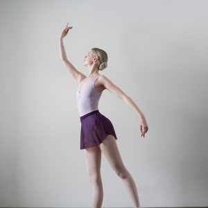 Op de afbeelding: Een jonge vrouw in een balletpose, gekleed in een lichtpaarse maillot en een donkerpaarse tule rok. Ze draagt roze balletten en heeft haar haar in een knotje.