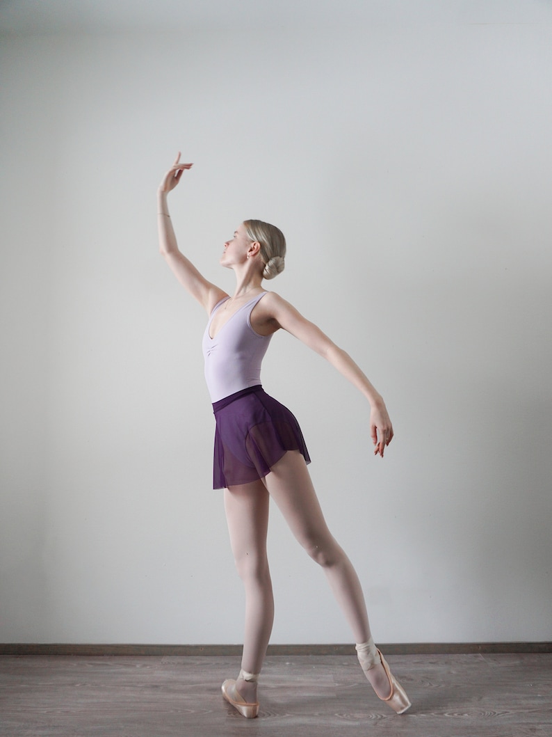 Op de afbeelding: Een jonge vrouw in een balletpose, gekleed in een lichtpaarse maillot en een donkerpaarse tule rok. Ze draagt roze balletten en heeft haar haar in een knotje.