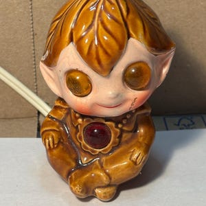 May include: A vintage ceramic figurine of a seated elf-like creature. The figurine has large, round eyes, a red gem, and a leaf-shaped head covering. The color palette includes shades of brown, tan, and pink.